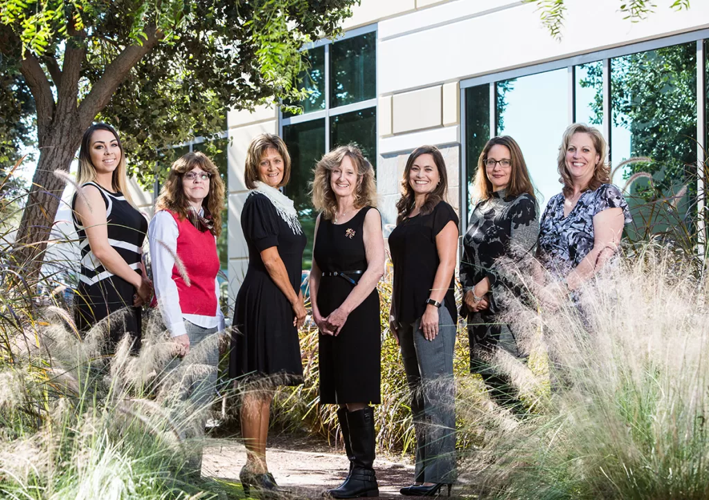 Seven women standing outside in front of a building with large windows and surrounded by greenery. They are posing for a group photo, smiling, and dressed in casual to semi-formal attire. Tall grass and trees frame the scene.