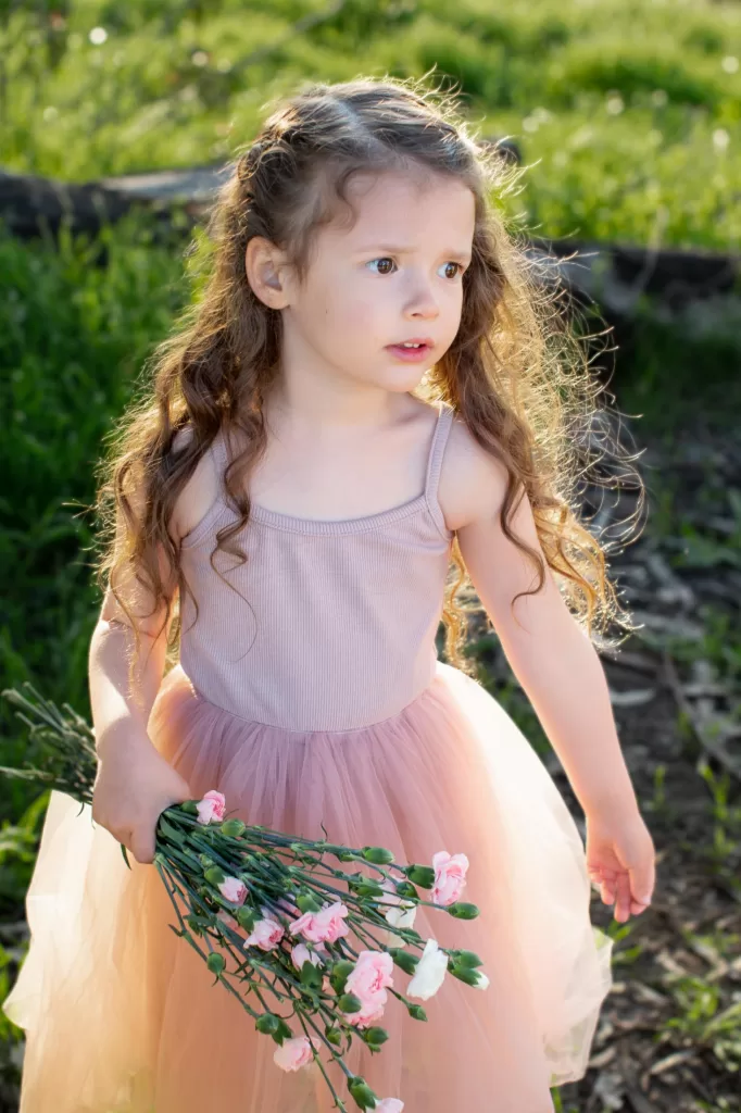 mother's day session showing A young girl with long, curly hair stands outdoors in a sunlit area, wearing a sleeveless pink dress with a tulle skirt. She holds a small bouquet of pink flowers in one hand and looks to the side with a thoughtful expression. Grass and trees are visible in the background.