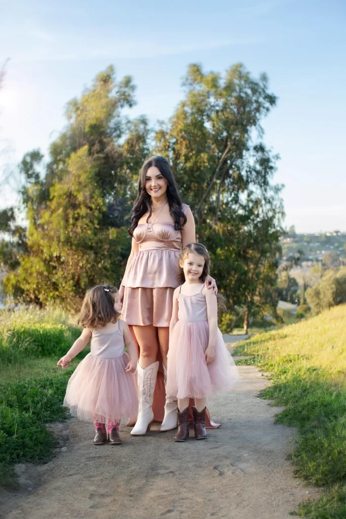 mother's day session showing a woman in a pink dress stands on a path flanked by two young girls in matching pink tutu dresses. One of the girls is looking up at the woman, while the other is walking away. They are outdoors with greenery and trees in the background.