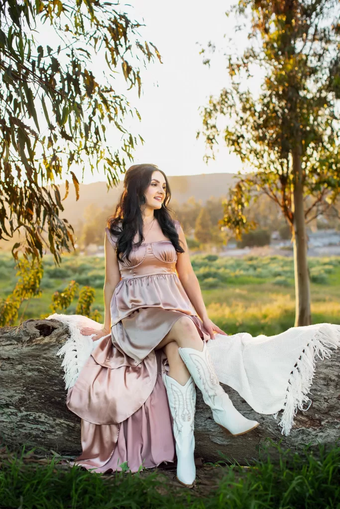 mother's day session showing a woman with long dark hair is sitting on a fallen tree trunk outdoors, draped in a white shawl. She's wearing a pink flowing dress and white cowboy boots. The background features sunlit trees, grass, and distant hills, creating a serene natural setting.
