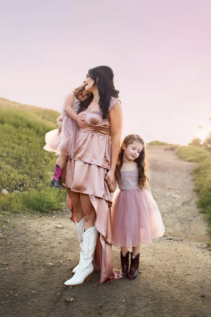mother's day session showing A woman in a silky pink dress and white cowboy boots stands on a dirt path, holding a small child in her arms while another child clutches her hand. They are surrounded by green fields under a soft, pink sky—a perfect moment for any family photographer.
