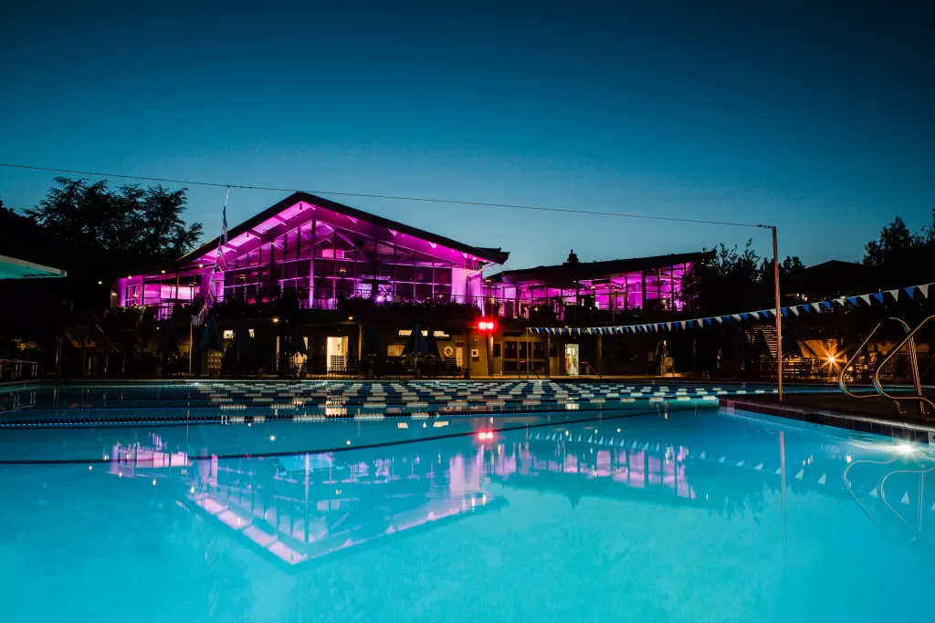 A brightly lit building with glass walls glows in purple illumination against the evening sky, setting a magical scene for a Palo Alto Bat Mitzvah. In the foreground is a swimming pool with clear blue water, reflecting the vibrant lights from the building. Trees and other structures surround the serene setting.