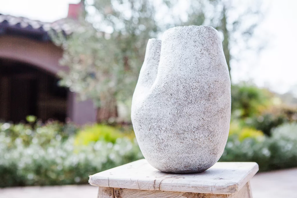 A white, abstract stone sculpture with smooth, curving surfaces sits on a wooden pedestal outdoors. In the background, there is a blurred view of greenery and a house.