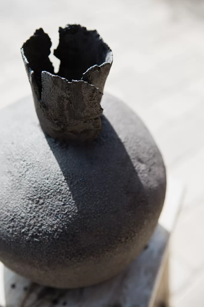 A weathered, round black sculpture with a jagged, cylindrical opening at the top, sits on a stone base. The surface appears rough and textured, and sunlight casts a shadow on one side. The background is blurred, highlighting the prominent features of the sculpture.