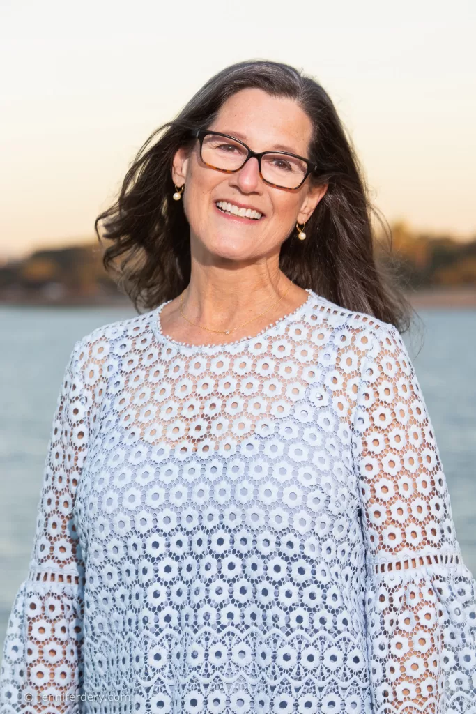 A woman with long brown hair, wearing glasses and a light blue, patterned blouse, stands smiling in front of a body of water with a blurred background of trees and sky at sunset.