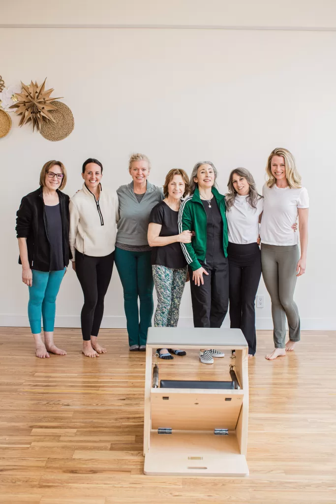 A group of seven women standing in a pilates studio, smiling. They are dressed in athletic wear and standing on a wooden floor. There is a Pilates reformer machine in front of them and wall decorations on the left.