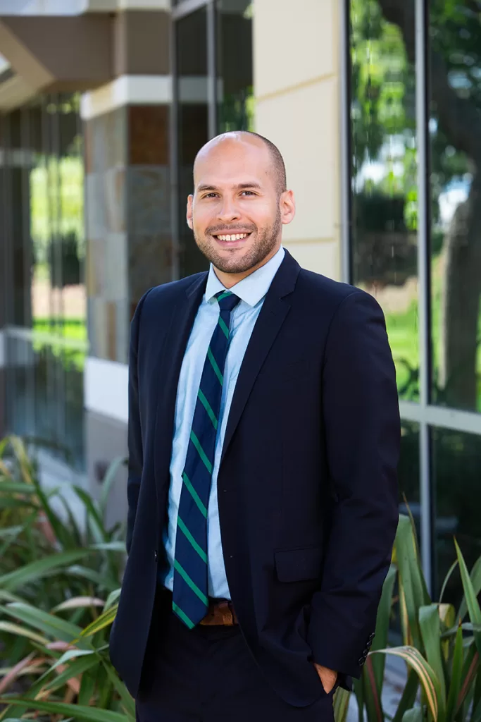 A smiling man with a shaved head and a beard wearing a navy blue suit with a blue and green striped tie stands in front of a modern building with large windows. Green plants with long leaves are in the foreground.