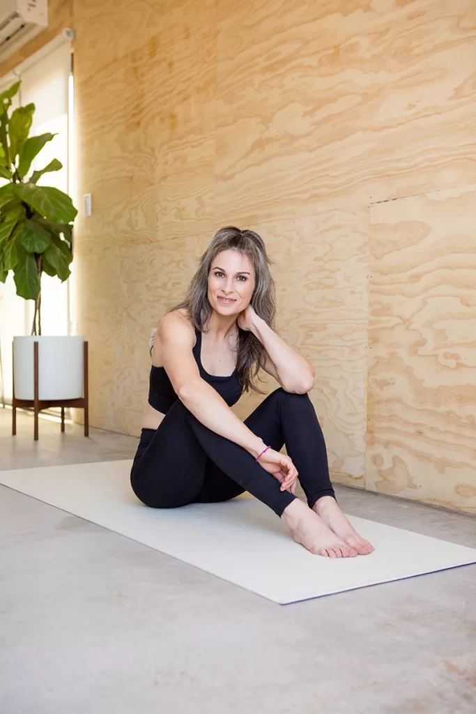A woman with long hair, dressed in black workout clothes, sits on a white pilates mat in a light, minimalistic room. She is smiling, with one hand resting on her neck and the other bent, supporting her seated pose. A large potted plant is in the background.
