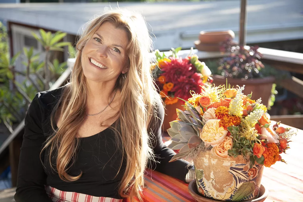 A smiling woman with long, blonde hair sits outdoors next to a vibrant floral arrangement in a decorative vase. She is wearing a black top and a striped apron. The background features greenery and outdoor furniture, creating a warm and inviting atmosphere.