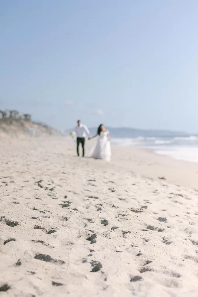 A bride and groom walk hand in hand on a sandy beach with the ocean and hills in the background, capturing the quintessential essence of wedding photography. The image is softly focused, creating a dreamy, ethereal effect. The bride wears a white dress, and the groom is dressed in a white shirt and dark pants.