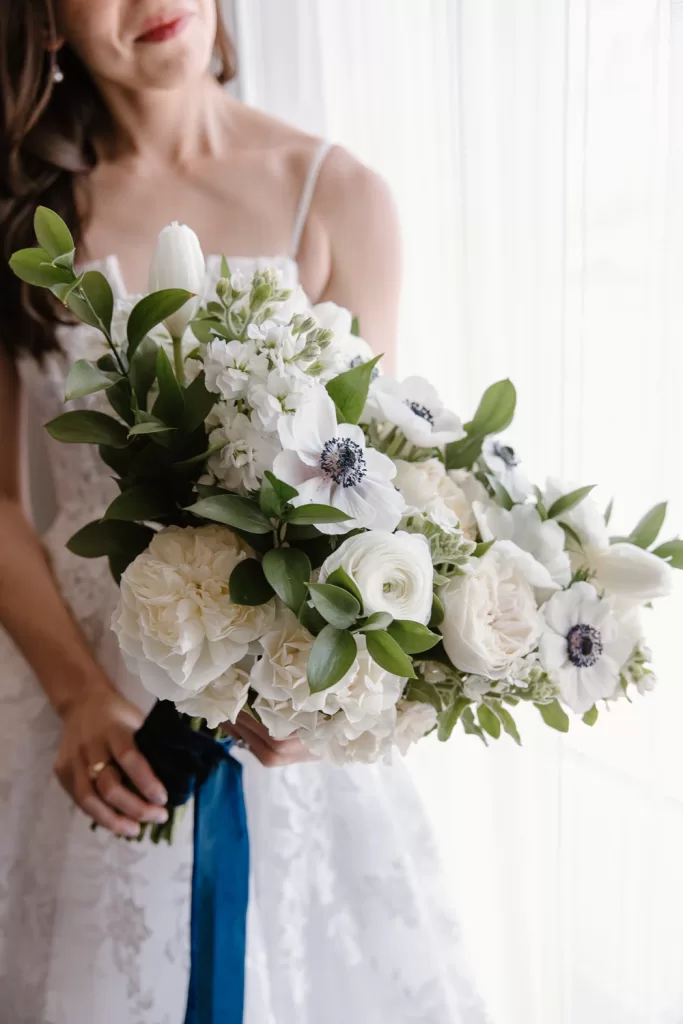 A bride stands gracefully in her white lace dress, holding a bouquet of white roses and anemones wrapped with dark blue ribbon. Behind her, the sheer curtain softly diffuses the light, creating an elegant ambiance reminiscent of a celebration at Southpointe Ballroom.
