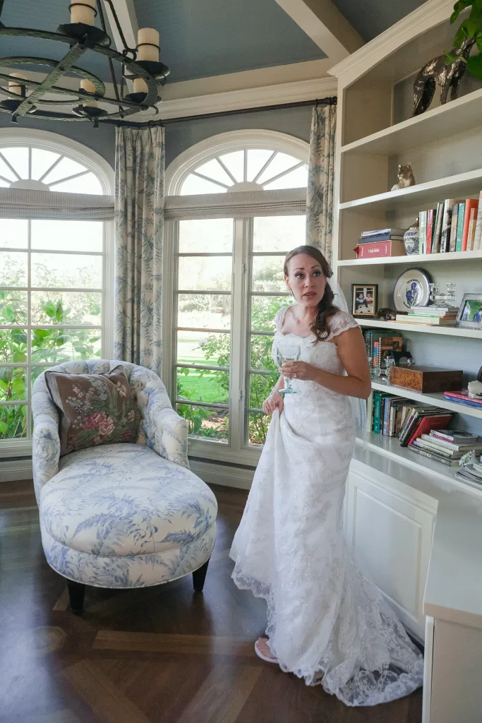 A bride in a white lace gown stands in a room with tall arched windows, holding a glass. The room features built-in bookshelves filled with books and decor, and a cushioned chair with a floral pattern. Natural light fills the space, highlighting the wooden floor.