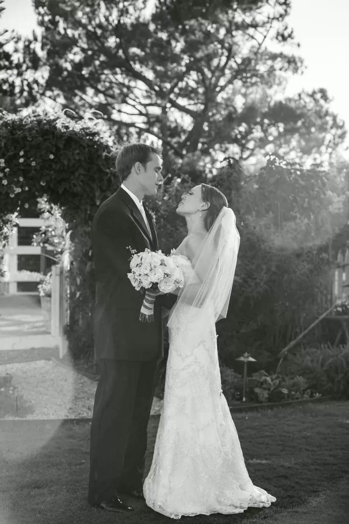 Black and white photo of a bride and groom standing outdoors on grass, facing each other and holding hands. The bride is wearing a flowing gown and veil, holding a bouquet of flowers. Trees and garden arch are visible in the background.