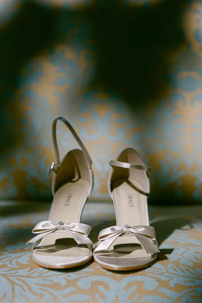 A pair of elegant beige high-heeled shoes with ankle straps and decorative bows on the toes are displayed against a patterned backdrop with soft blue and gold tones.