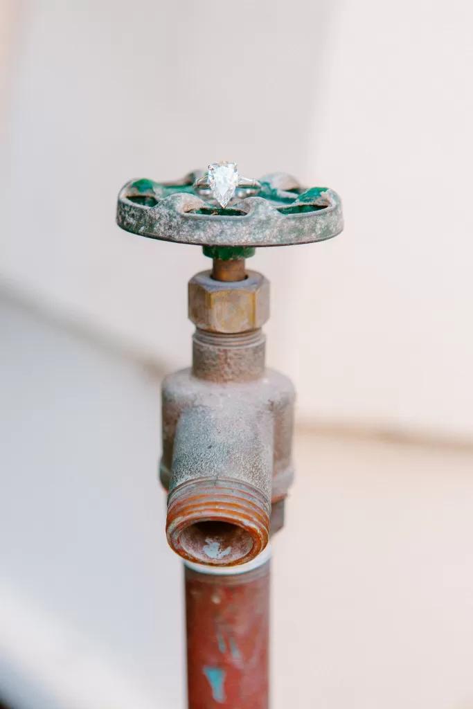A close-up image of a rusted outdoor water valve with a pear-shaped engagement ring balanced on top. The ring features a large central gemstone, and the valve handle displays signs of green and brown patina. The background is blurred and neutral-toned.