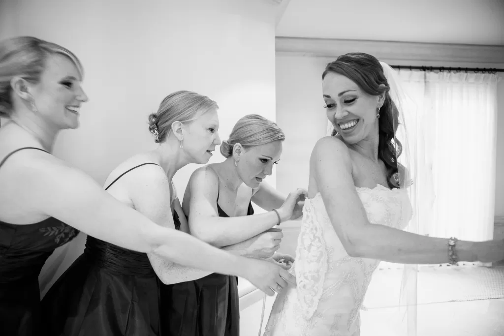 A black-and-white photo shows a smiling bride in her wedding dress being helped by three bridesmaids. The bridesmaids are adjusting the back of the bride's dress while sharing a joyful moment. The scene appears to be in a well-lit room.