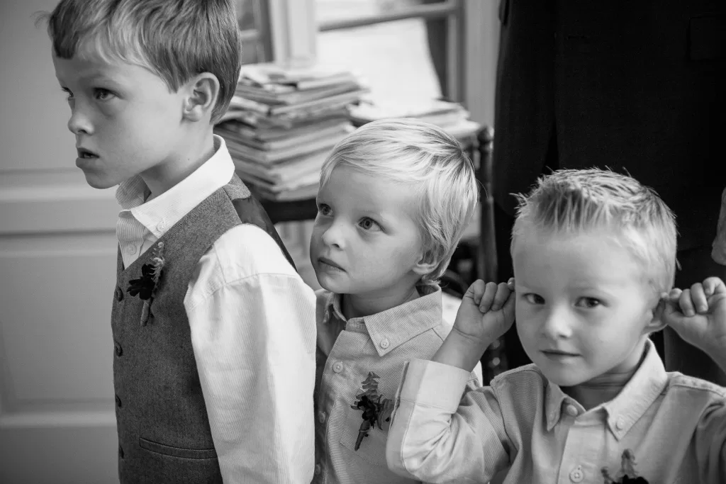 A black and white photo of three young boys standing in a room. They are all dressed in button-up shirts, and one has a vest on. The boy in the center looks curious, the one on the left appears thoughtful, and the boy on the right is pulling on his ears playfully.