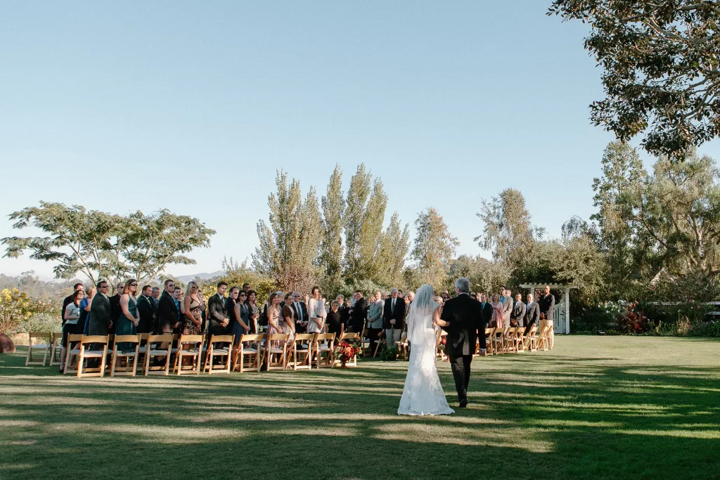 A bride, wearing a white wedding dress and veil, walks down an outdoor grassy aisle accompanied by an older man in a suit. Guests seated on wooden chairs on either side of the aisle turn to watch them. Trees and a clear blue sky form the background.