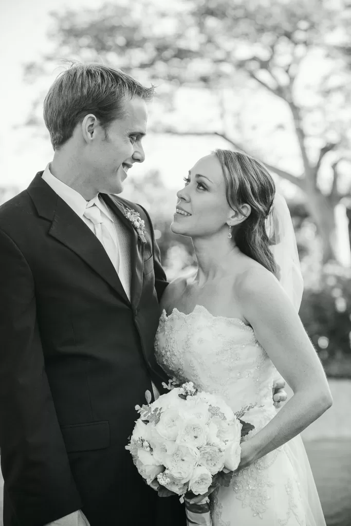 A bride and groom gaze into each other's eyes, smiling softly. The bride holds a bouquet of flowers and wears a strapless gown and veil, while the groom dons a suit and tie. They stand outdoors, with blurred trees in the background, creating a serene backdrop.