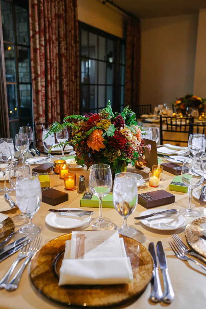 An elegant dining table set for a formal event, featuring gold-rimmed plates, folded napkins, and menus. The centerpiece is a vibrant floral arrangement surrounded by small lit candles. The setting is illuminated by natural light coming through large windows in the background.