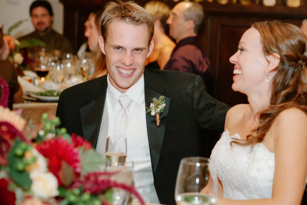 A groom in a black suit and white tie sits next to a bride in a white wedding dress. Both are smiling and laughing. They are seated at a decorated table with glasses and floral arrangements, and other guests can be seen in the background.