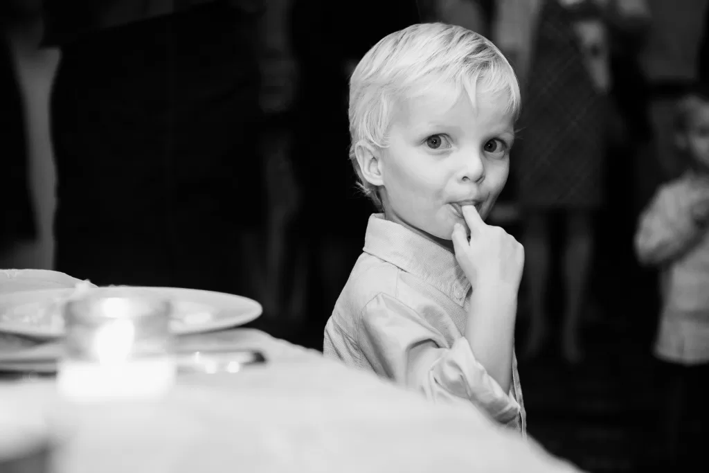 A young child with blonde hair looks towards the camera while sucking on their finger. The child is wearing a light-colored shirt and standing next to a table with plates and candles. The background shows other people, slightly out of focus. The image is in black and white.