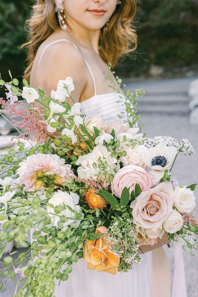A woman in a white dress holds a large bouquet of assorted flowers, including roses, greenery, and small white blooms. The background is outdoors with greenery.