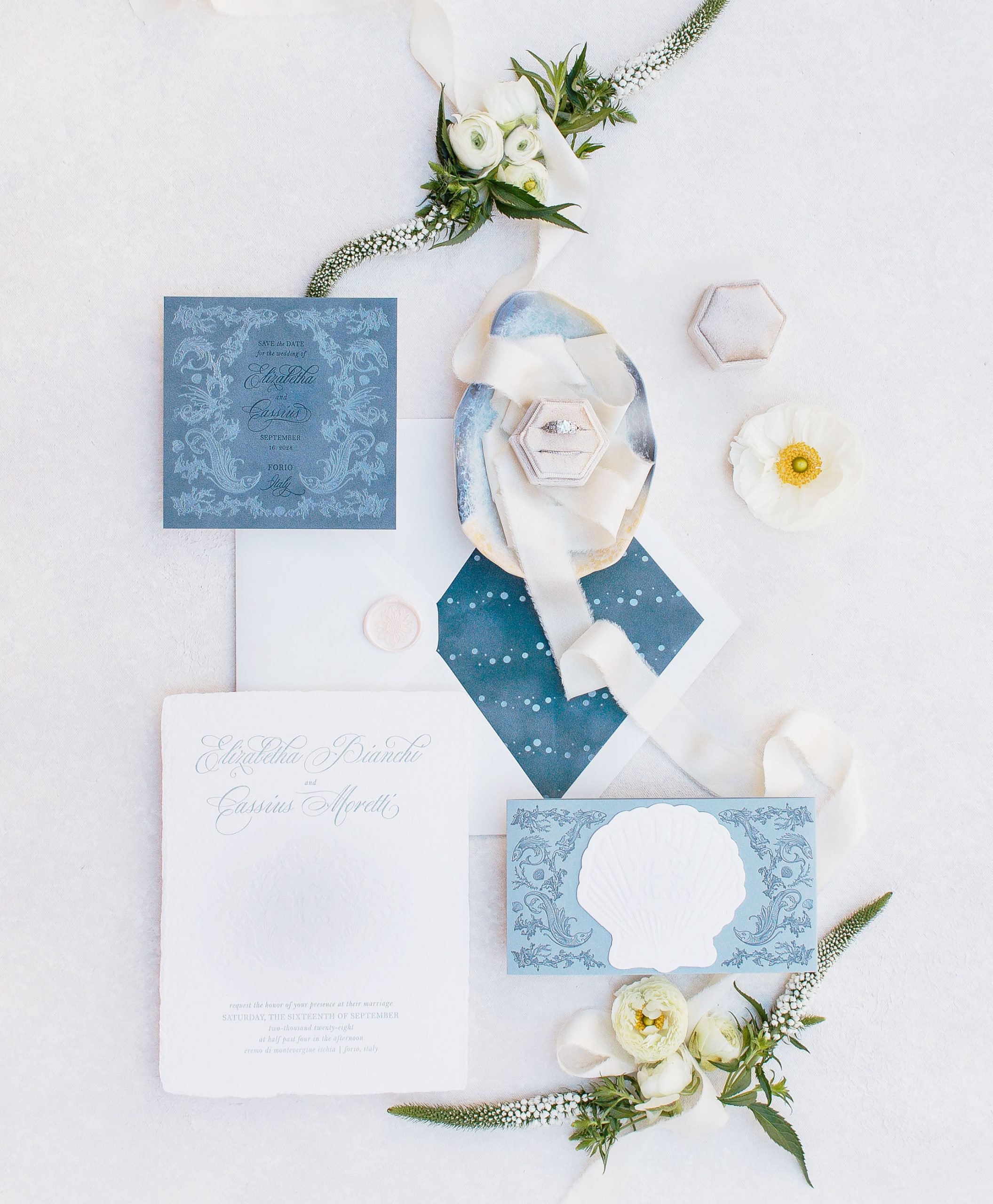A flat lay of elegant blue and white wedding invitations with script text, decorative patterns, and a seashell motif. Surrounded by white flowers, greenery, and two small boxes, one containing a ring.