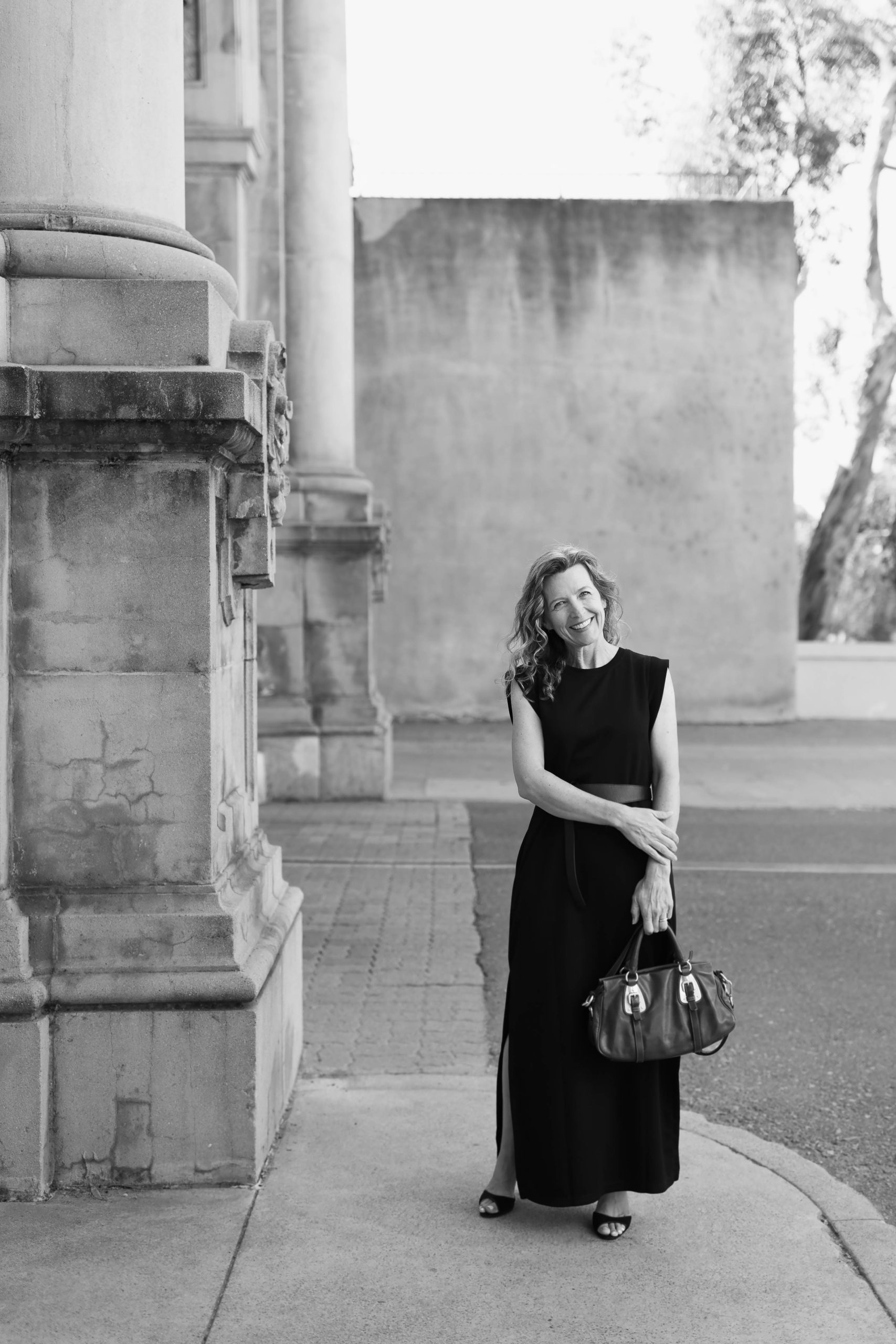 Photographer Jennifer Dery in a black dress stands smiling on a sidewalk next to a large stone column. She is holding a handbag, and the background shows a building with textured walls and leafy trees. The image is in black and white.