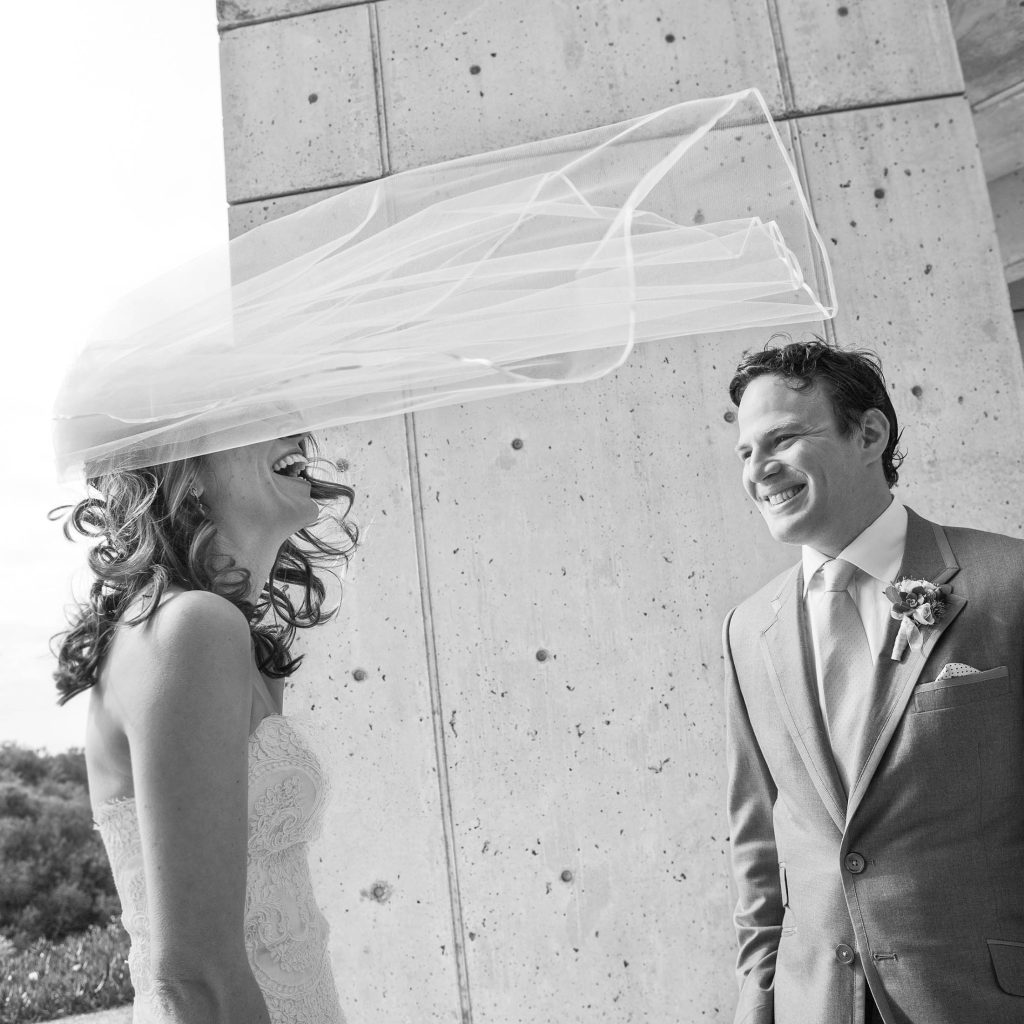 A black and white photo of a bride and groom laughing. The bride's veil is caught in the wind, flowing above her. They are standing outside in front of a concrete wall. The groom is wearing a suit with a boutonniere.