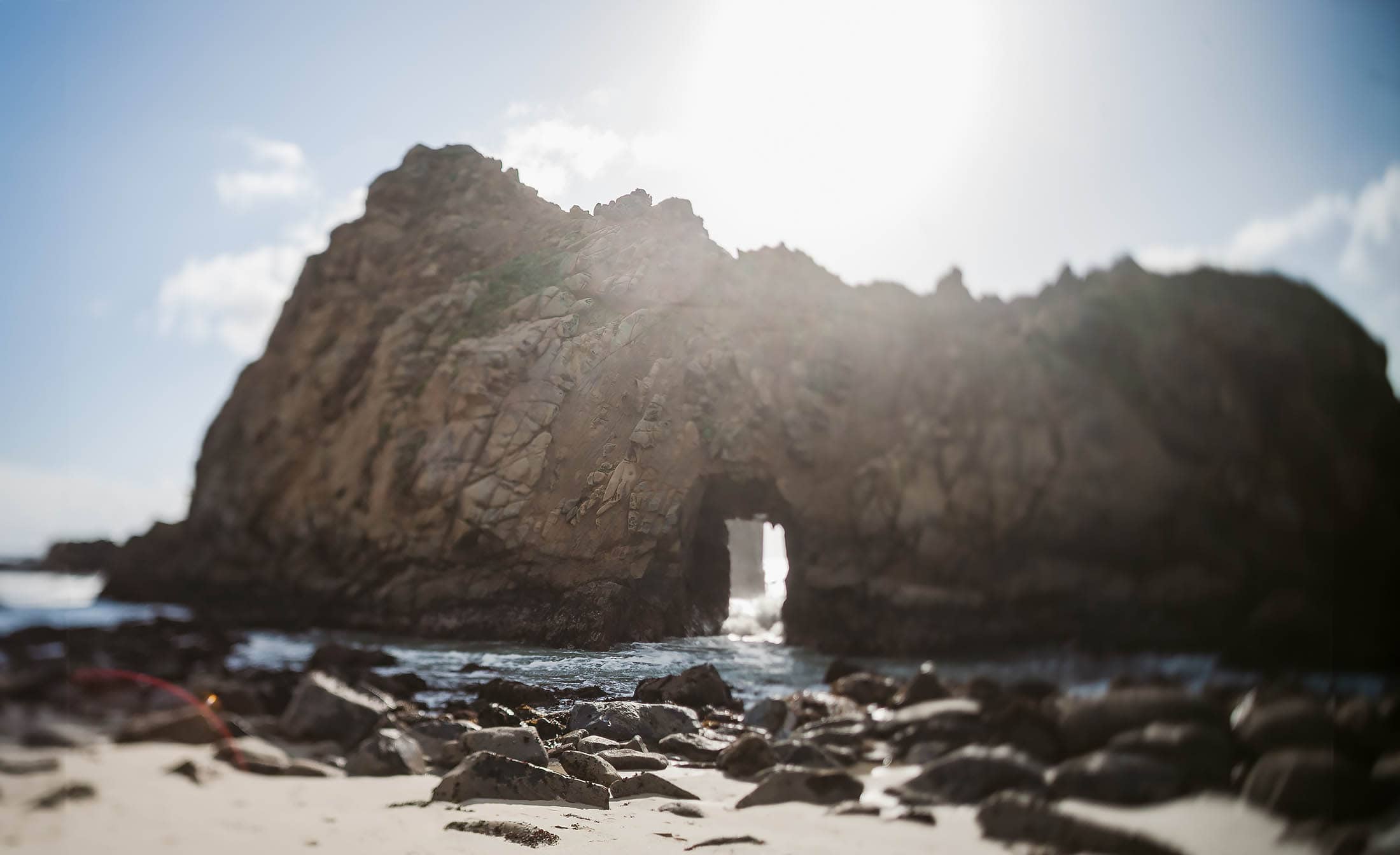 A large rock formation with an archway stands by the ocean under a bright sun. Waves lap against the rocky shoreline, and the sandy foreground is scattered with boulders. The sky is mostly clear, with a few clouds.