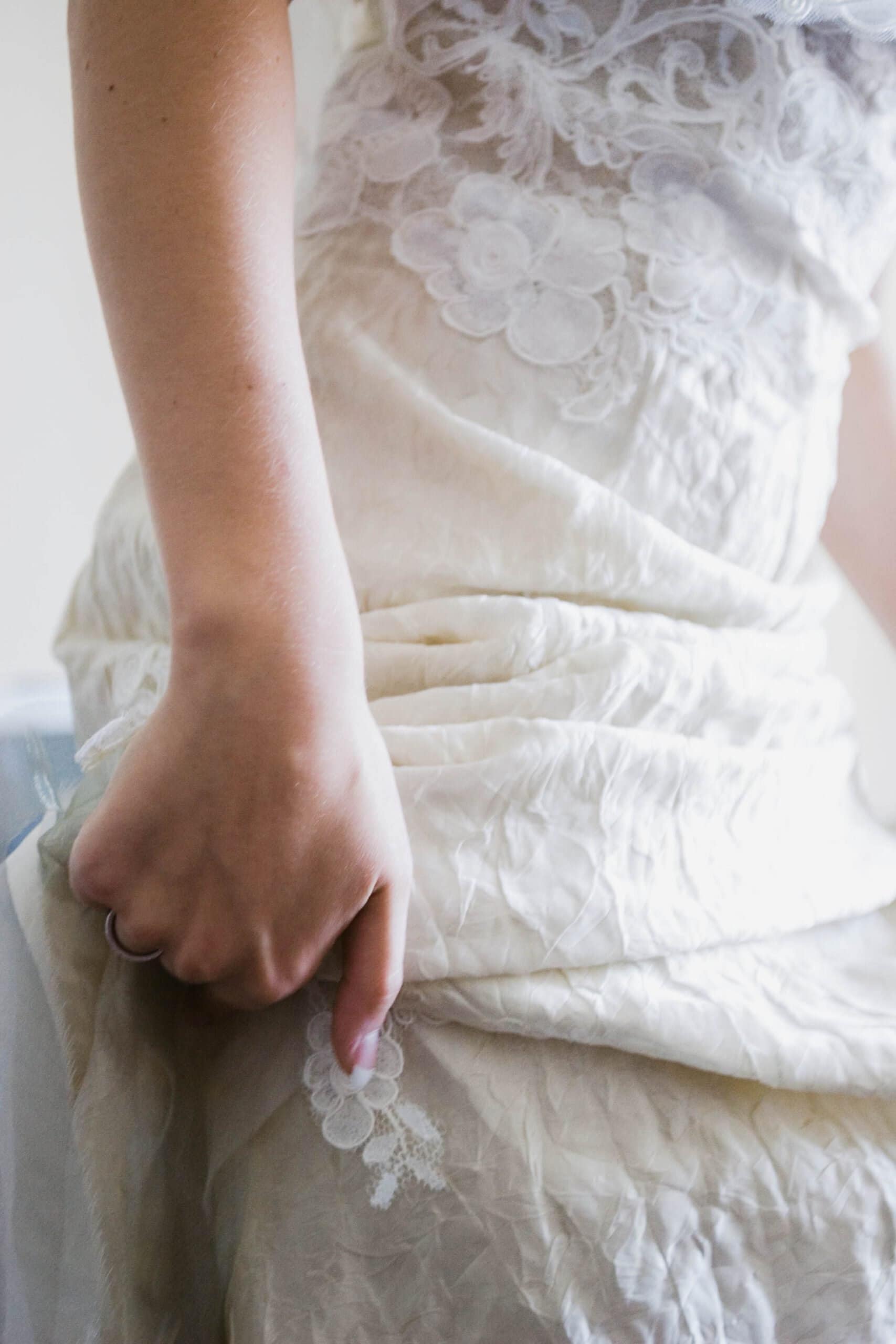Close-up of a person in a crinkled, embroidered white lace dress, gently holding the fabric with their hand, wearing a ring. The dress features floral patterns near the top and a soft, flowing texture.