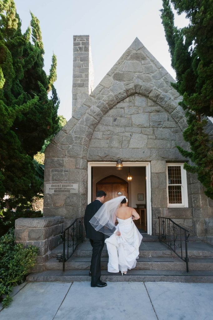 A bride and groom are walking up the steps to a stone chapel surrounded by tall trees. The bride is wearing a white dress and veil, and the groom is in a black suit. They are heading towards the entrance.
