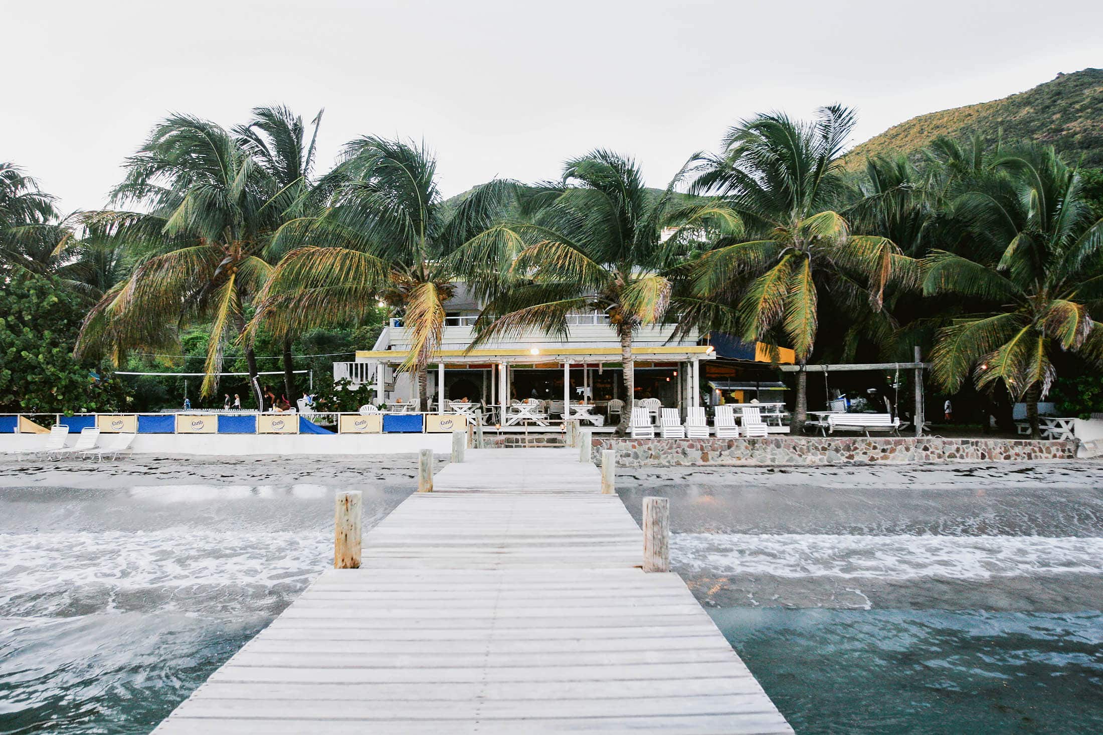 A wooden pier leads to a beachside restaurant surrounded by tall palm trees. There are tables and chairs on a deck, and gentle waves reach the sandy shore. A hill is visible behind the trees under a clear sky.