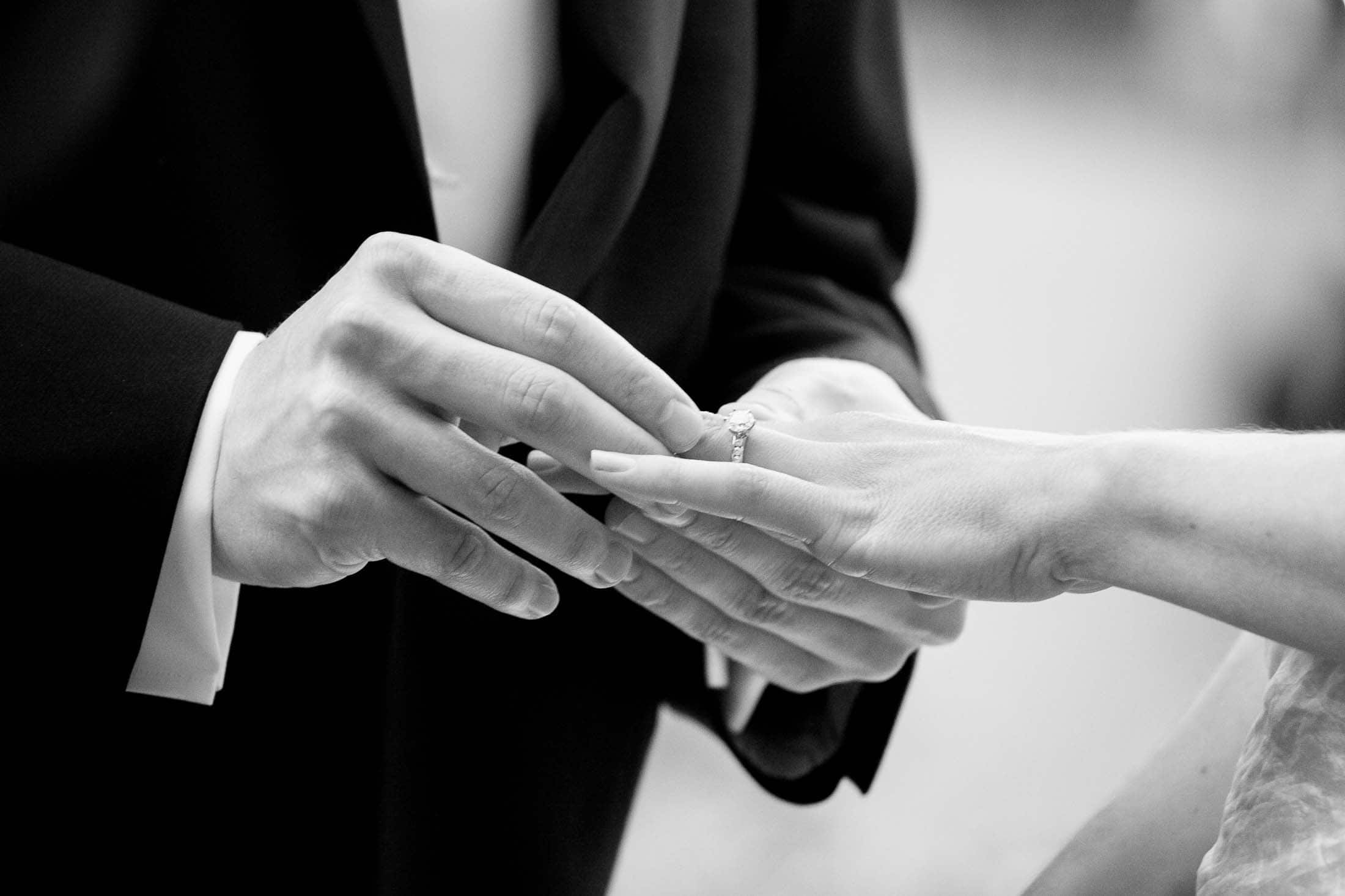 A black and white photo of a close-up scene where one person is placing a ring on another person's finger. The individuals are dressed formally, suggesting a wedding or engagement setting.