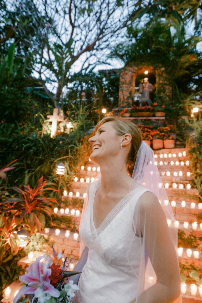 A smiling bride in a white dress and veil stands outdoors, holding a bouquet. Behind her, a staircase lined with glowing candles ascends through lush greenery, creating a romantic and serene atmosphere.