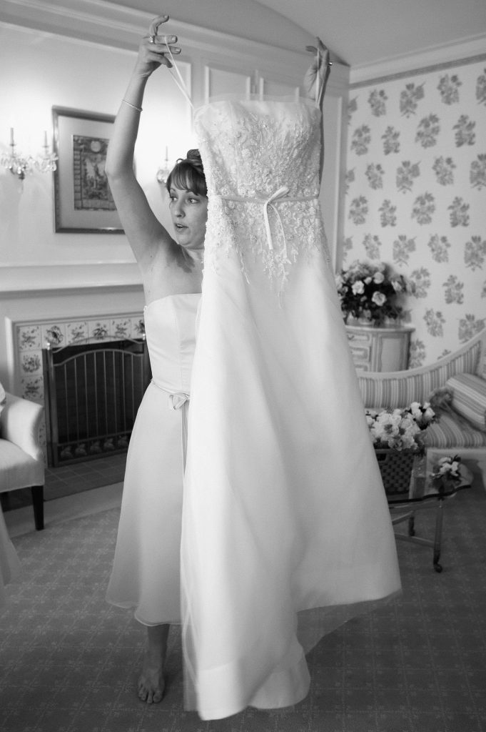 A woman holds up a wedding dress in a room decorated with floral wallpaper. She stands barefoot on a carpet, with furniture and a fireplace visible in the background. The dress has lace detailing. The scene appears to be a preparation for a wedding.