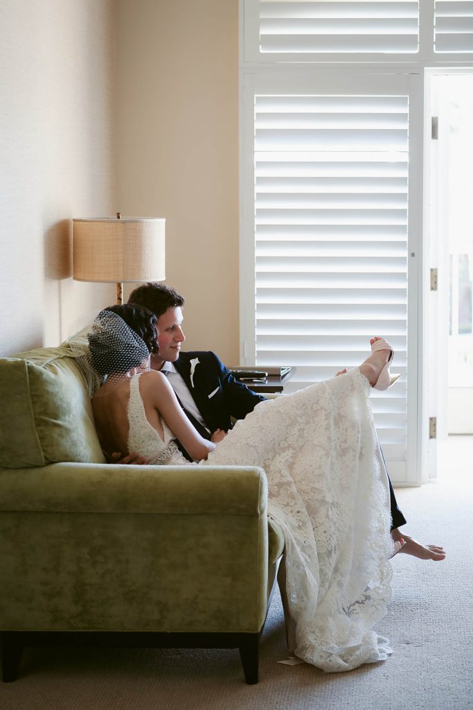A newlywed couple sits on a couch in a softly lit room. The bride wears a lace gown and leans against the groom, whose feet are propped up. They appear relaxed and content, next to a lamp and closed blinds.