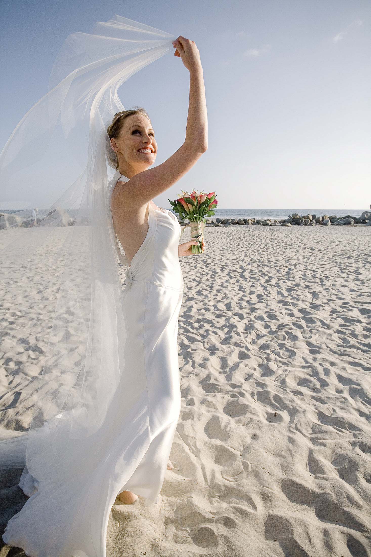 A bride in a flowing white gown and veil smiles and holds a bouquet of flowers on a sandy beach under a clear blue sky.