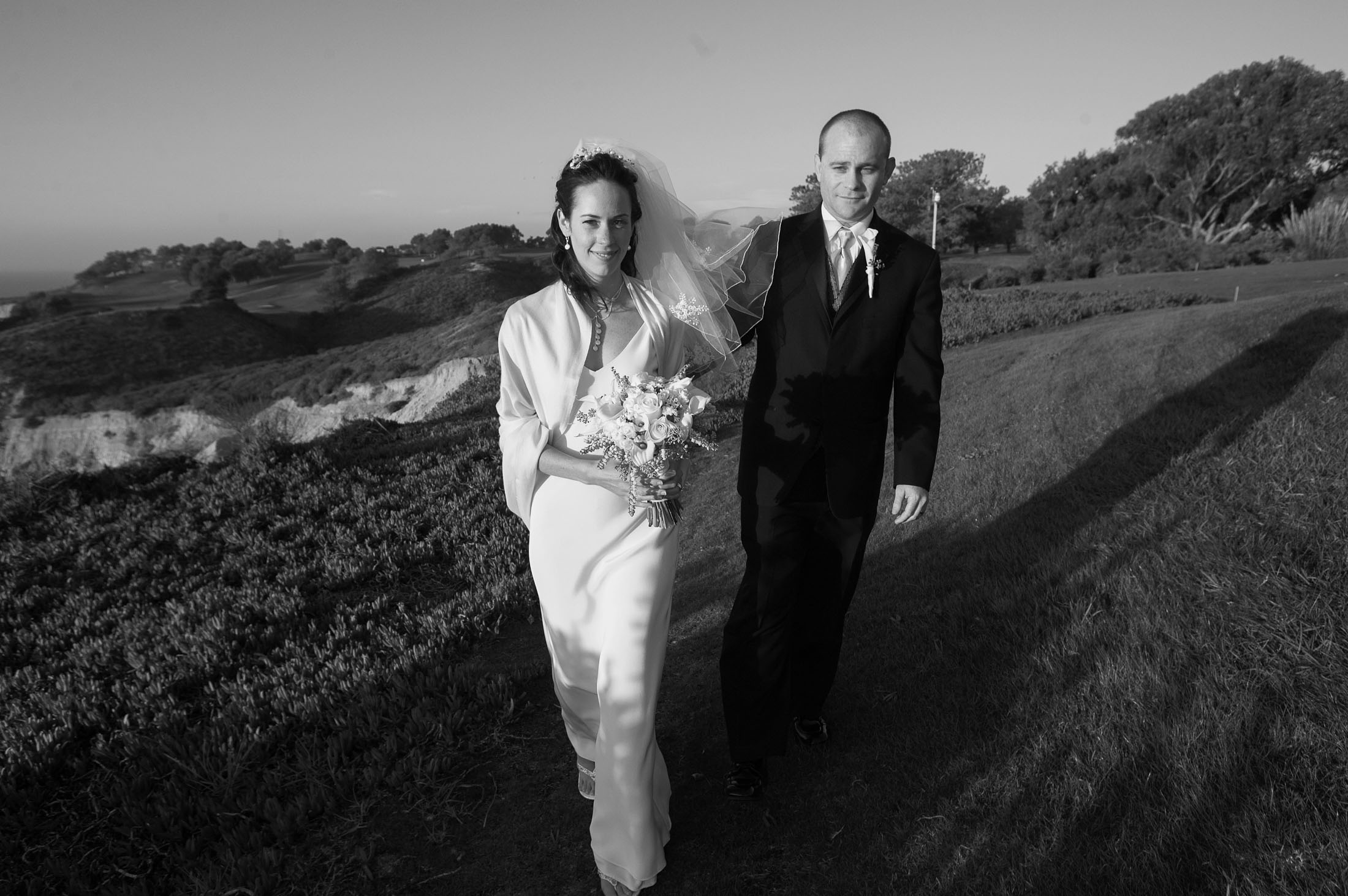 A bride and groom walk hand in hand on a grassy hillside. The bride holds a bouquet and wears a wedding gown with a veil, while the groom is in a suit and tie. The background features a scenic landscape with a clear sky. The image is in black and white.