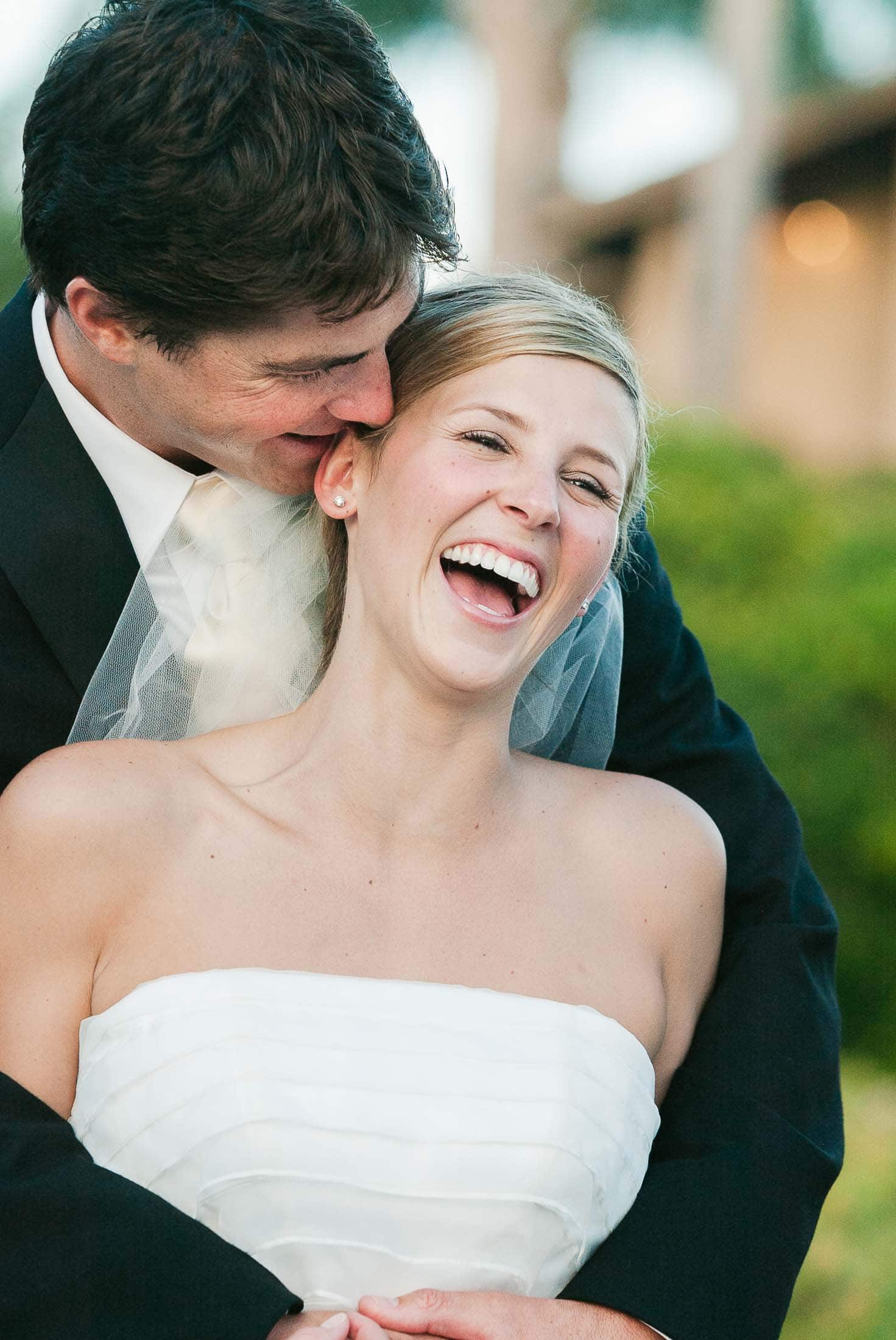A groom in a black suit embraces a laughing bride in a white strapless wedding dress. They are outdoors with blurred greenery and a building in the background. The bride is smiling widely, displaying joy and happiness.
