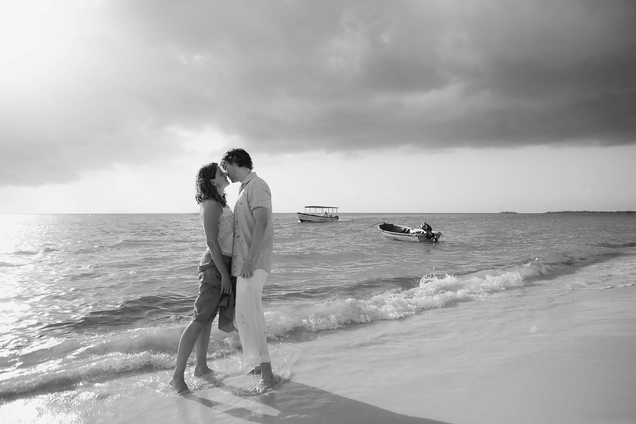 A couple stands on a sandy beach, embracing at the water's edge under an overcast sky. Two boats are visible in the calm sea. The scene is captured in black and white, creating a romantic and serene atmosphere.