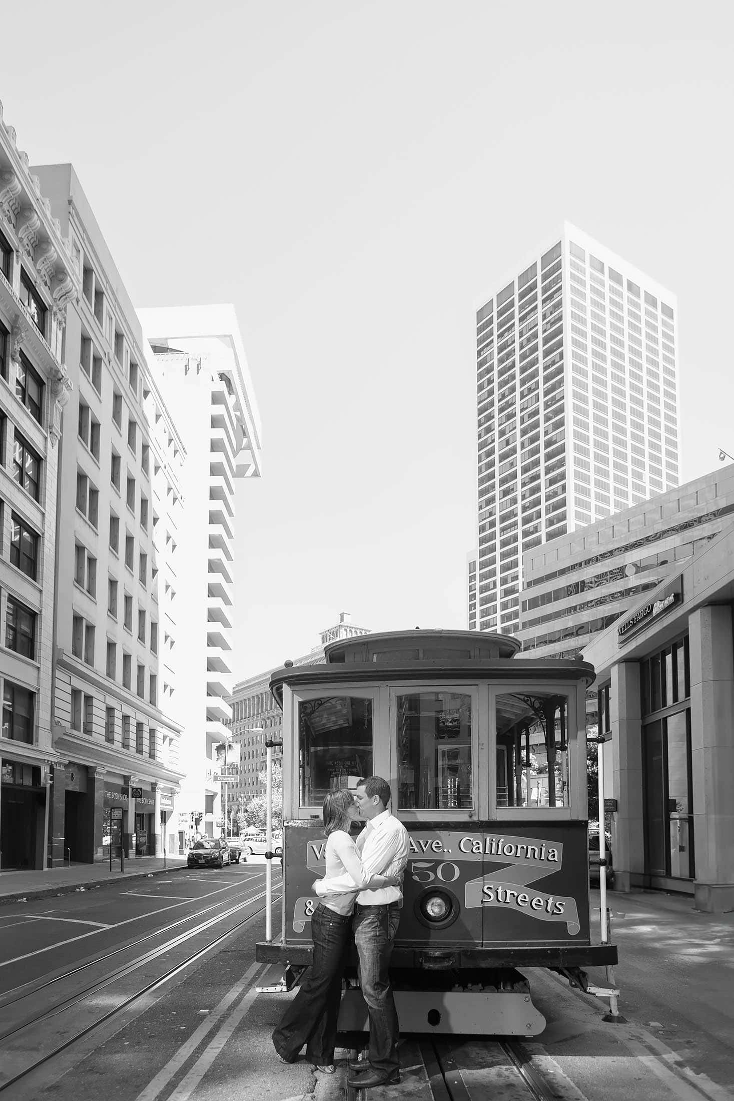 A couple kisses in front of a traditional San Francisco cable car on a sunny street. Tall buildings line the sides, and the cable car displays "California Streets" on its front. The scene captures a romantic moment in an urban setting.