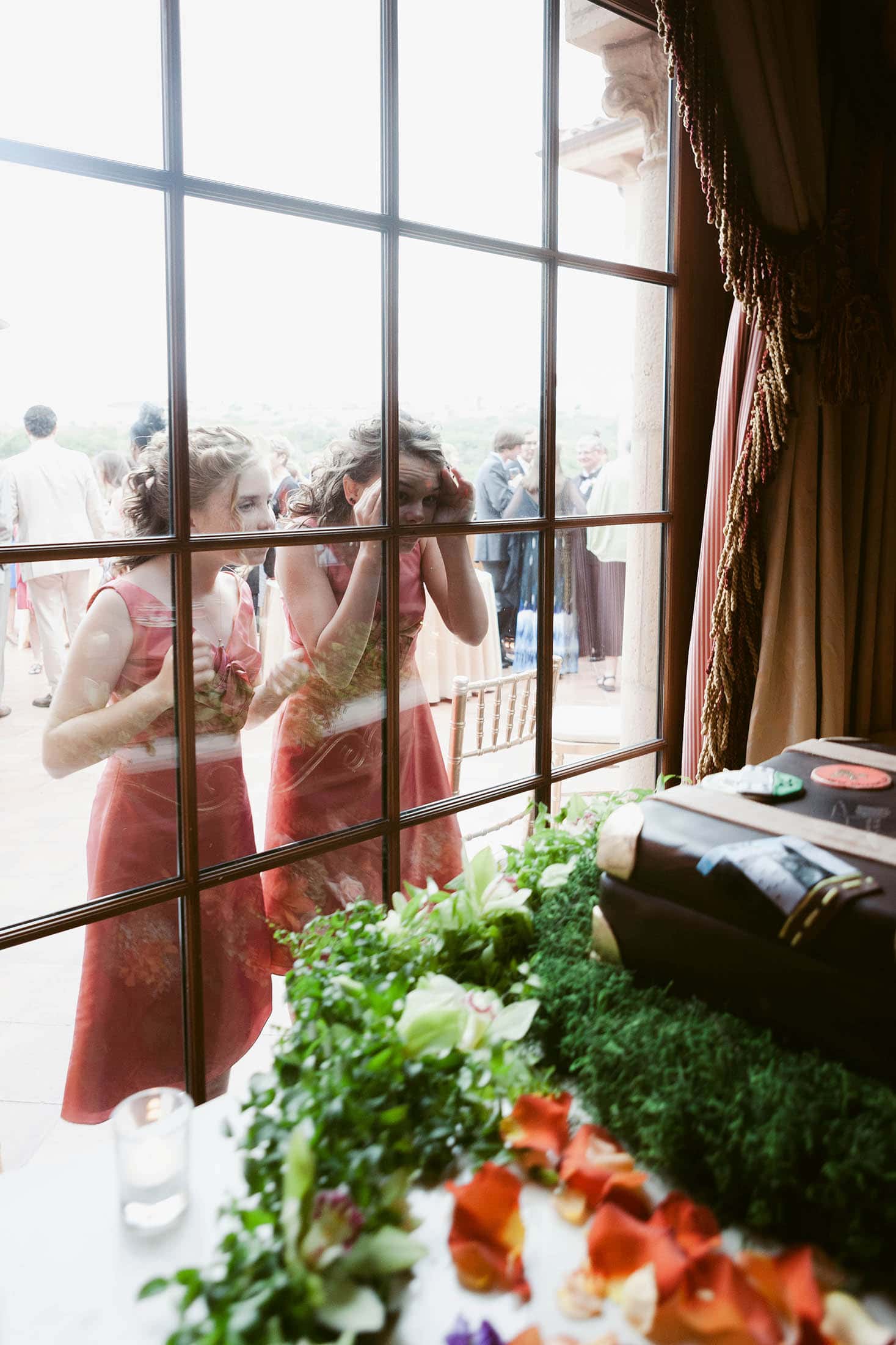 Two people in pink dresses look through a window from outside. They gaze at a table inside adorned with greenery and a decorative cake shaped like books. People are visible in the background.