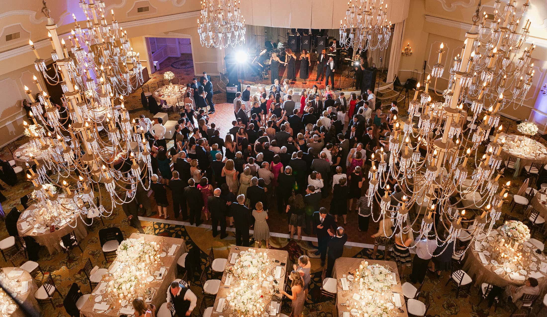 A large, elegant ballroom with numerous chandeliers overhead. Guests in formal attire gather around a stage where performers are entertaining. Tables are set with white tablecloths and floral centerpieces. The room is warmly lit.