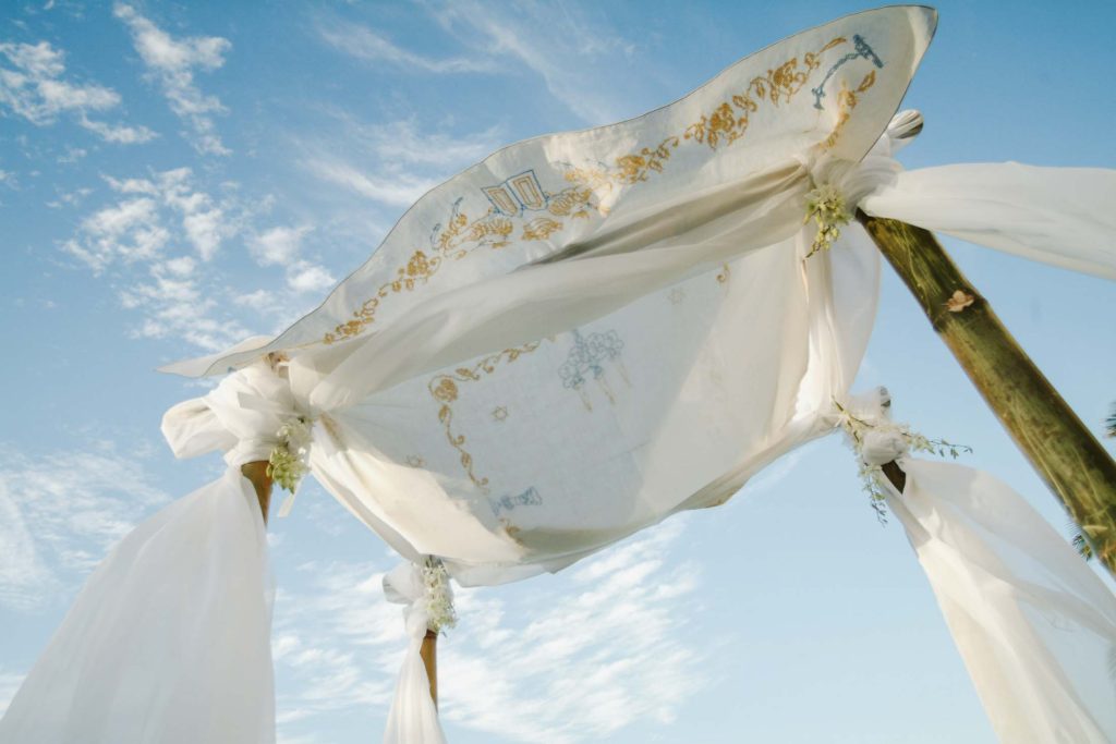 An outdoor chuppah with white fabric and gold embroidery is set against a blue sky with wispy clouds. The wooden posts are adorned with white cloth and small floral arrangements.