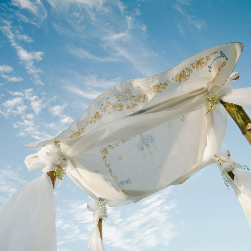 A white wedding chuppah stands against a bright blue sky with wispy clouds. The fabric covering is adorned with gold embroidery and delicate floral designs, draped over four wooden poles wrapped with white cloth and small floral decorations.