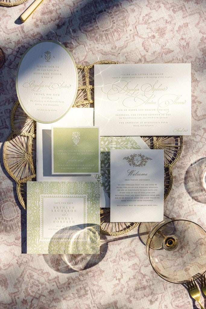 A collection of elegant wedding invitations and stationery set on a patterned surface. The papers feature delicate, intricate designs in shades of white and green, with cursive text. A shadow from a glass adds a touch of warmth to the arrangement.