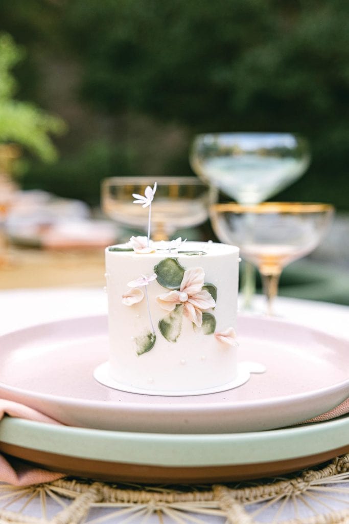 A small white cake decorated with pastel pink flowers and green leaves sits on a pale pink plate. The table setting includes elegant glassware with gold rims and layered plates, set outdoors in a garden-like environment.