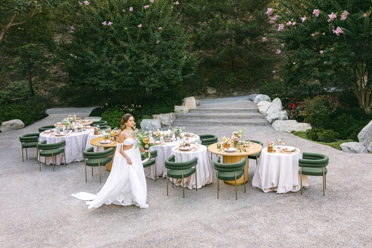A woman in a white dress walks past elegantly set tables with green chairs and floral centerpieces on a stone patio surrounded by greenery.